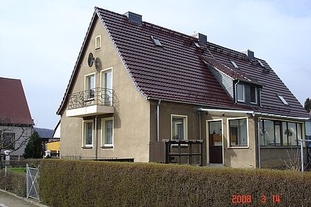 Ehemals weißes Einfamilienhaus mit Holzbalkon und ergrauter Fassade vor der Sanierung