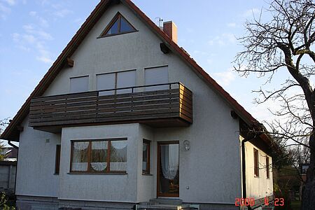 Ehemals weißes Einfamilienhaus mit angebautem Holzbalkon und ergrauter Fassade
