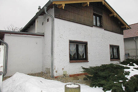 Weißes Wohnhaus mit bröckelnder Fassade im Erdgeschoss und Holzvertäfelung im oberen Stockwerk