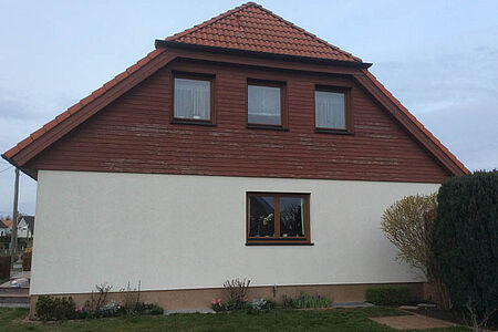 Zweistöckiges Wohnhaus mit schmutzig-weißer Fassade im Erdgeschoss und abgeschlagener Holzvertäfelung im oberen Stockwerk